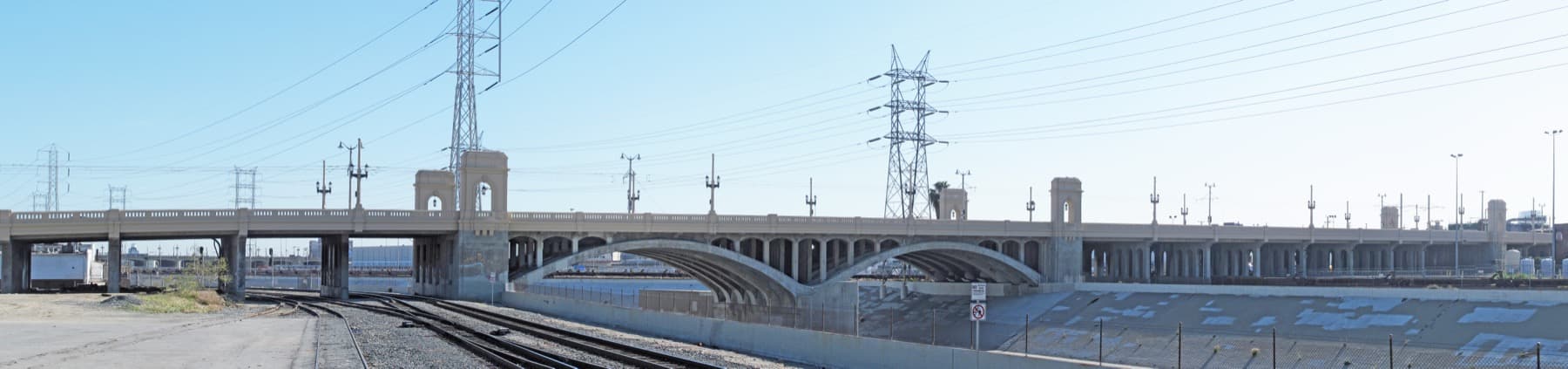 A long concrete bridge with arches and towers across a dry river channel and railway tracks