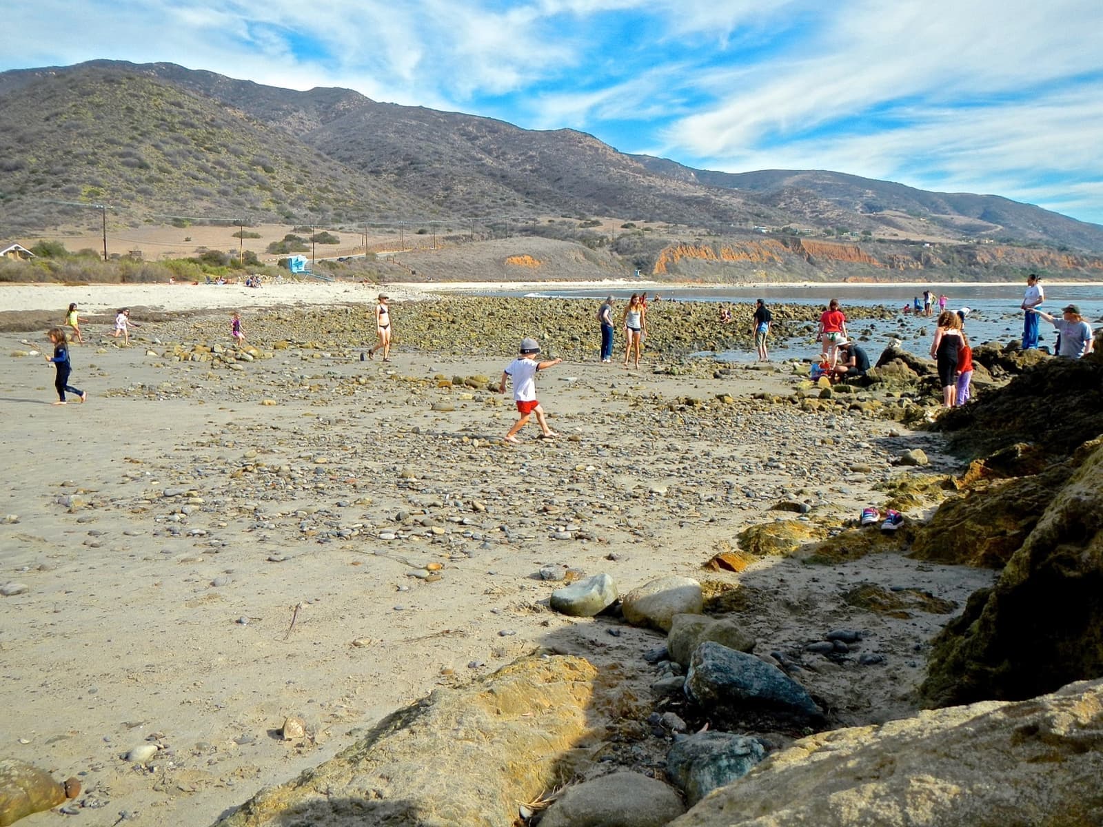A sunny beach with rocks and tide pools, people paddling and brown hills behind