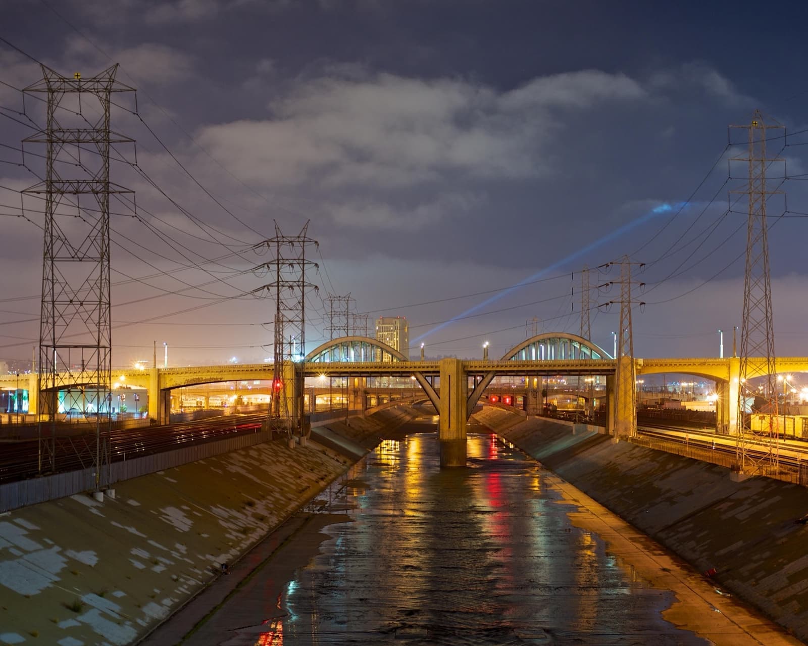 A lit steel arch bridge over a concrete river channel at dusk, reflections on the wet floor