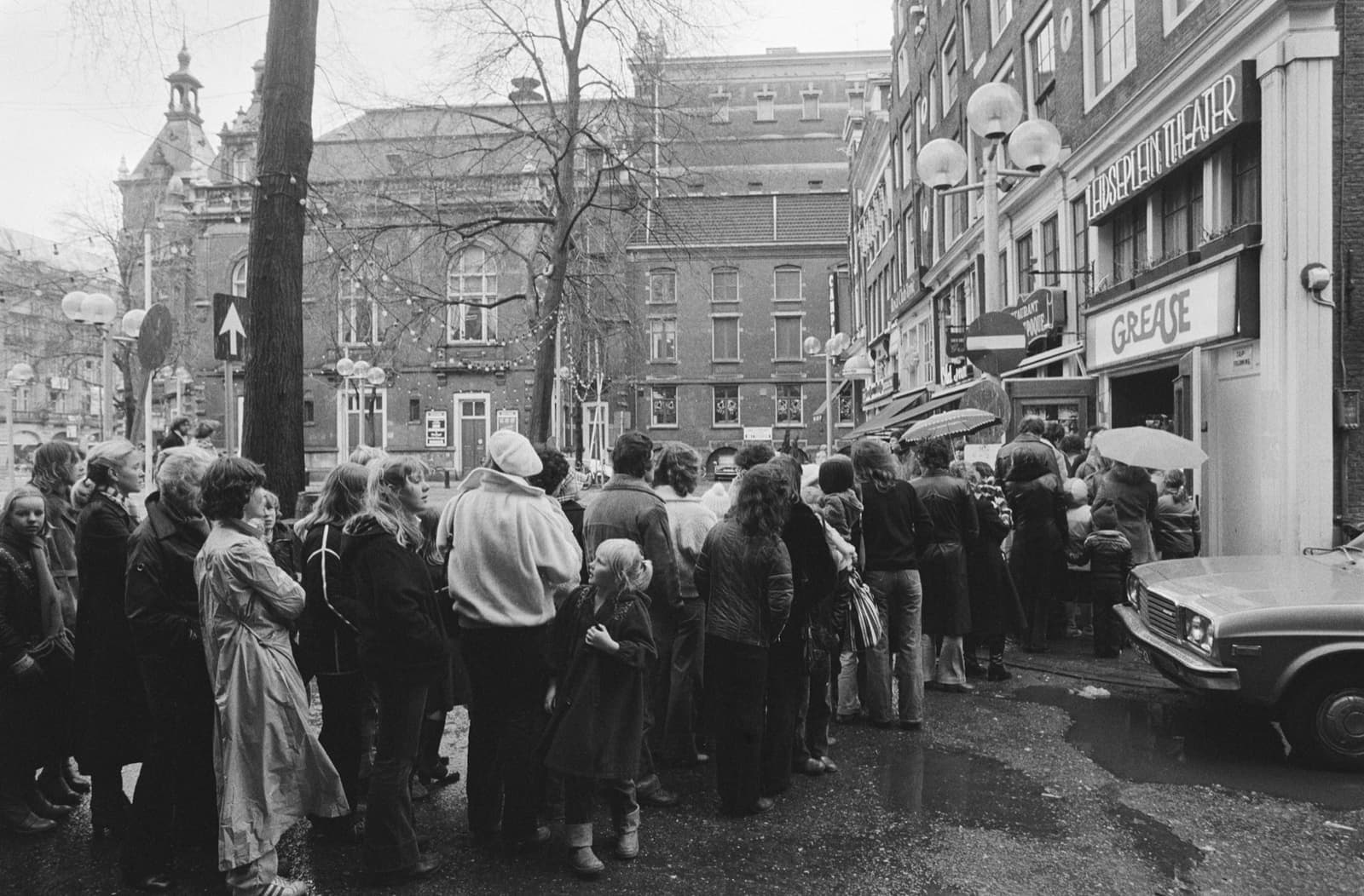 People in winter coats queue along a wet street beside a cinema with GREASE on its canopy