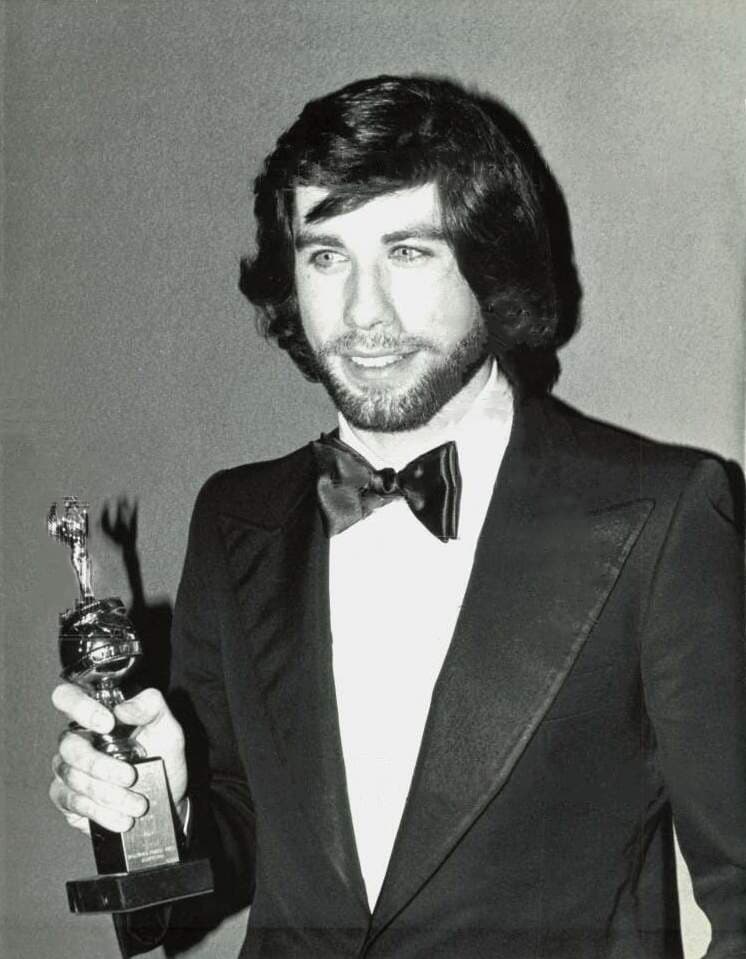 A young man with dark hair and a beard in a black tuxedo and bow tie, holding up a statuette