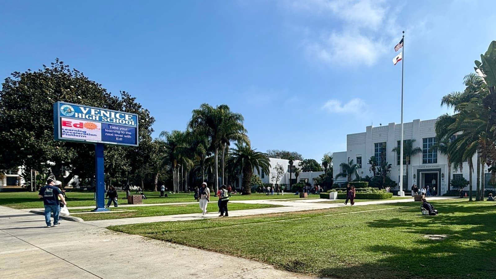 A white school building behind a lawn and palm trees, a blue Venice High School sign in front