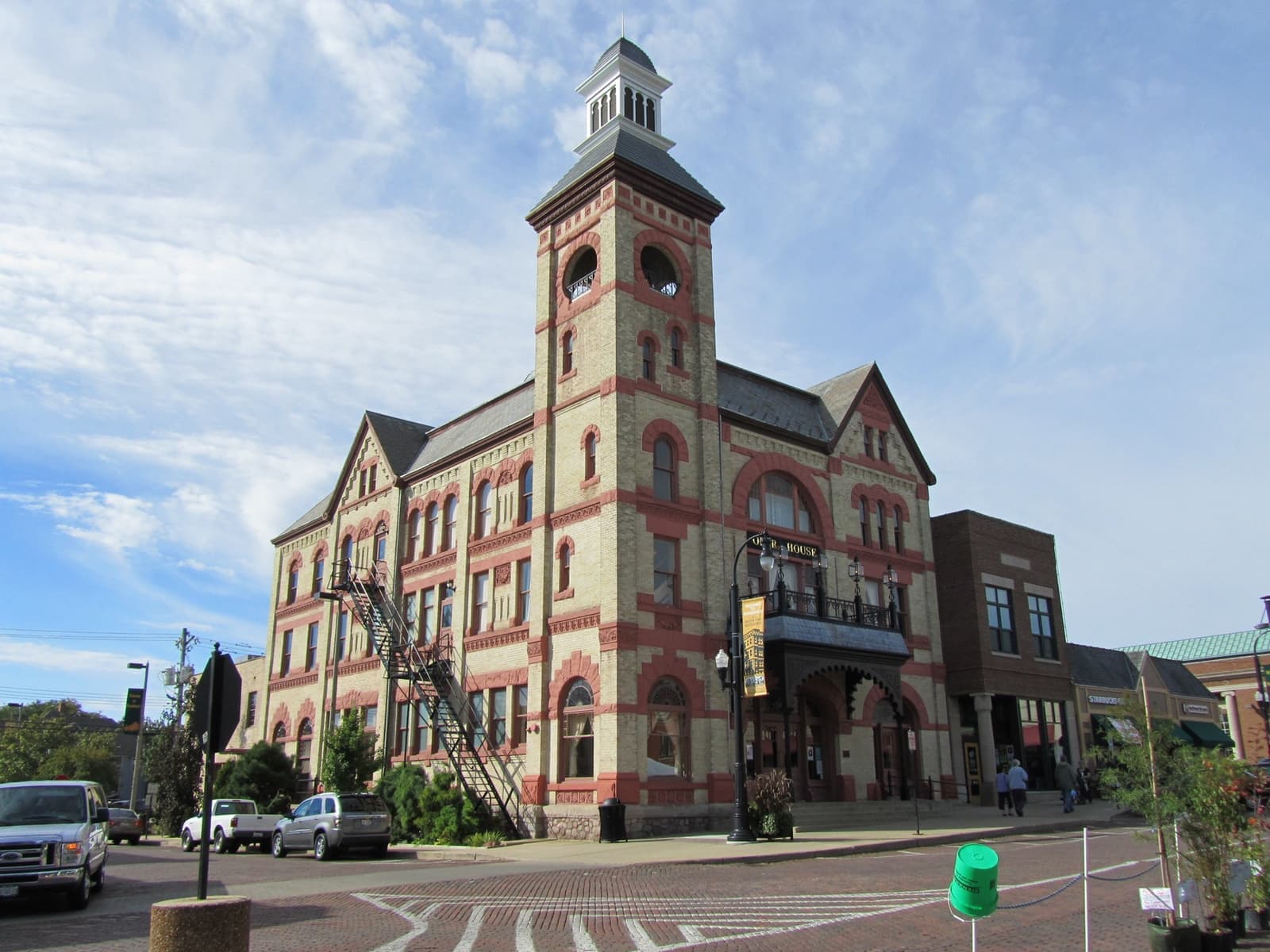 A tall yellow-brick Victorian building with a red-trimmed bell tower on a street corner