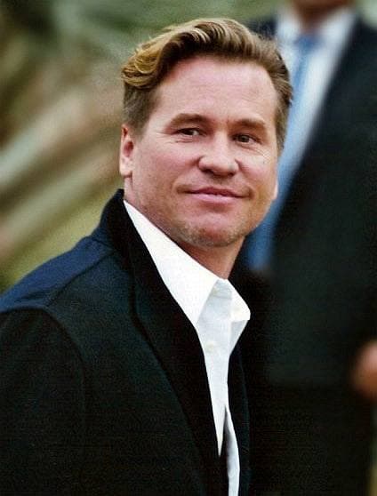 Val Kilmer in a dark jacket and open white shirt, smiling
