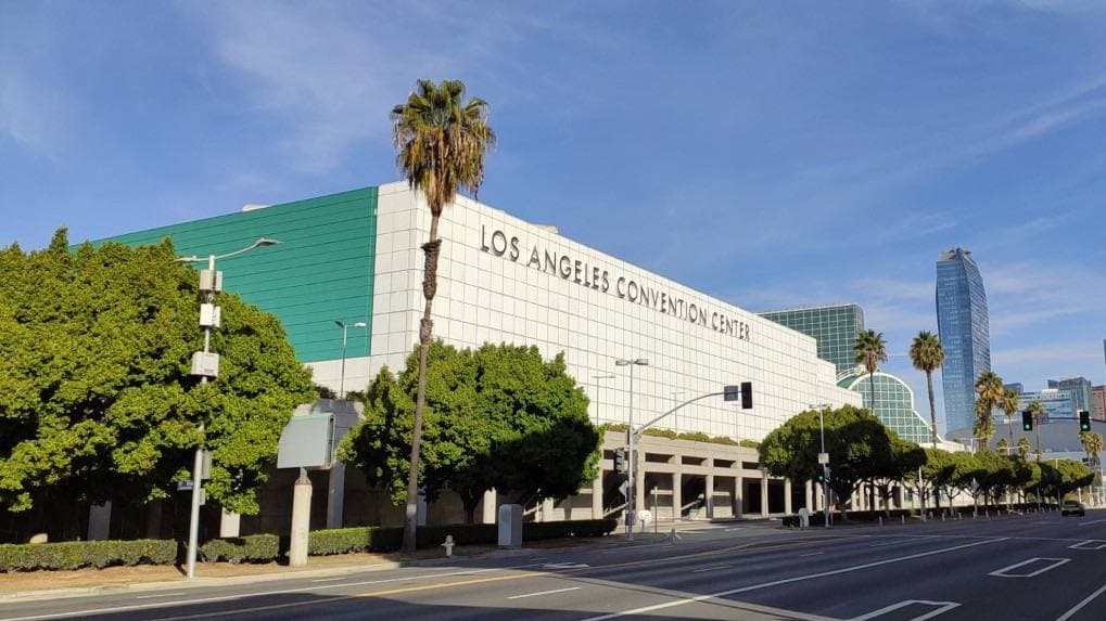 A long green-glass convention hall behind palm trees and a wide empty street