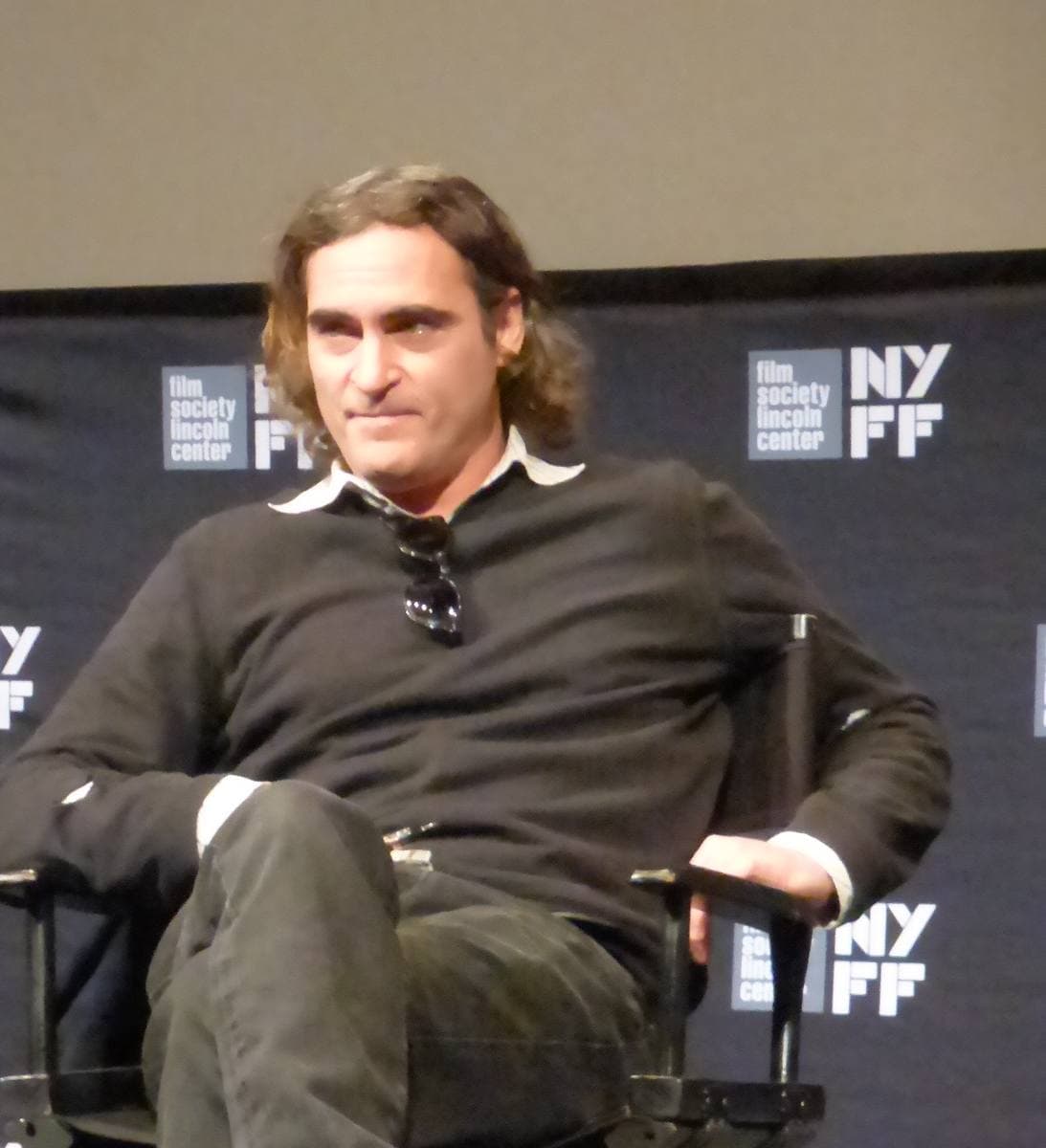 Joaquin Phoenix with shoulder-length hair and a dark jumper, seated, sunglasses hooked on his collar