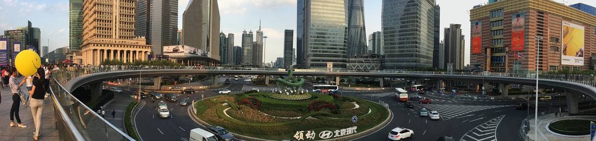 A wide panorama of a circular raised walkway above a round garden and traffic, with towers behind