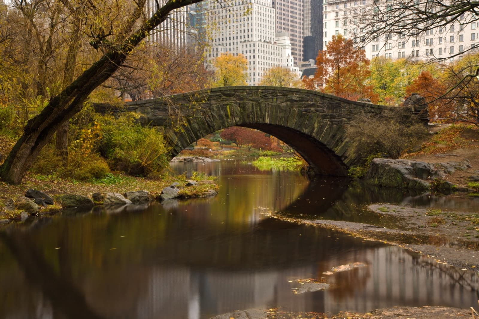 A stone arch bridge over still water among autumn trees, with towers behind