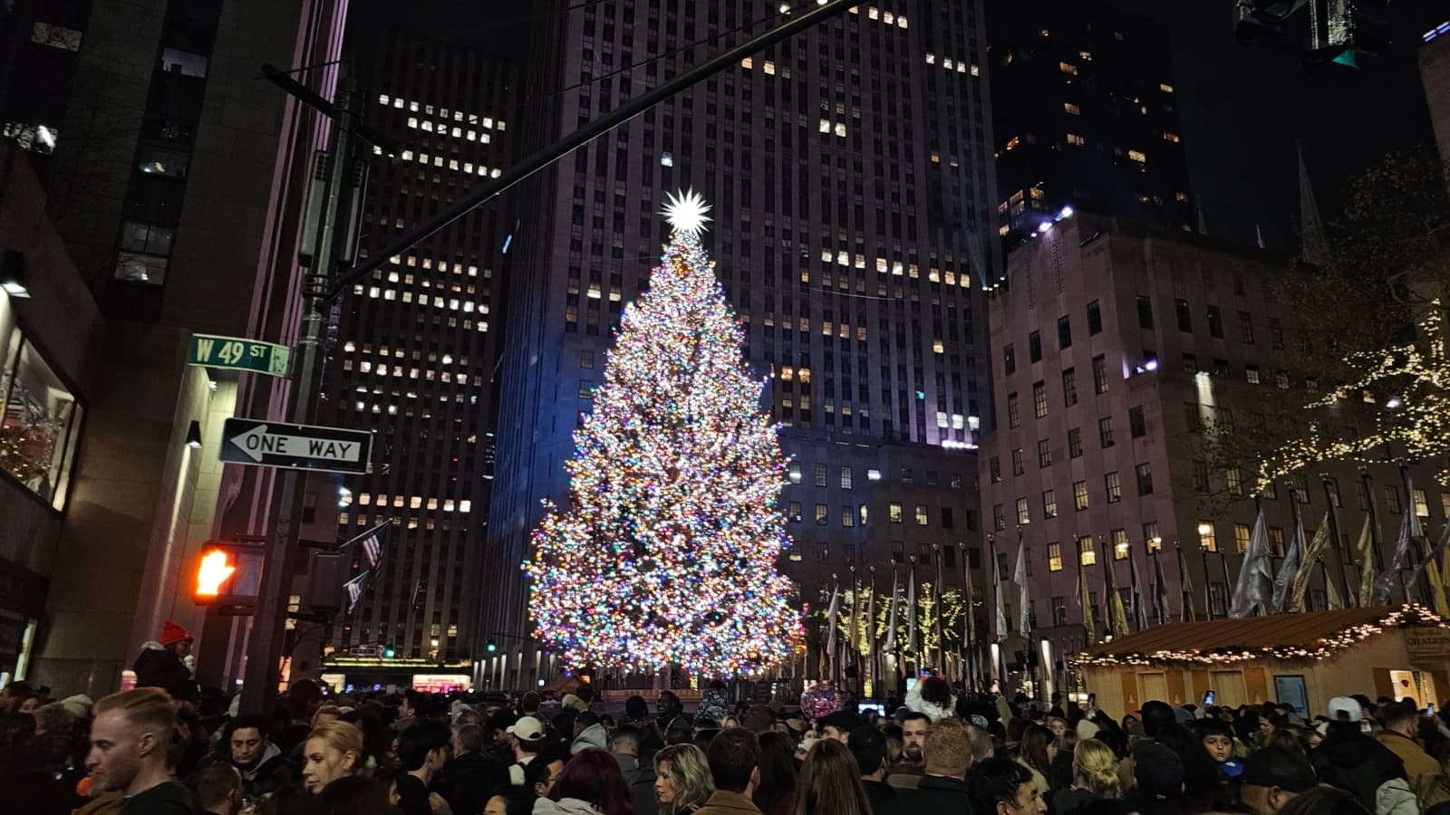 A lit Christmas tree among skyscrapers at night, with crowds below