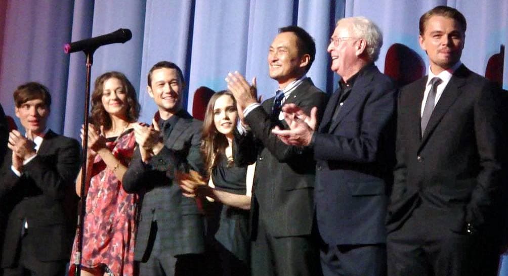 Seven actors in dark suits and dresses applauding on a stage in front of a curtain