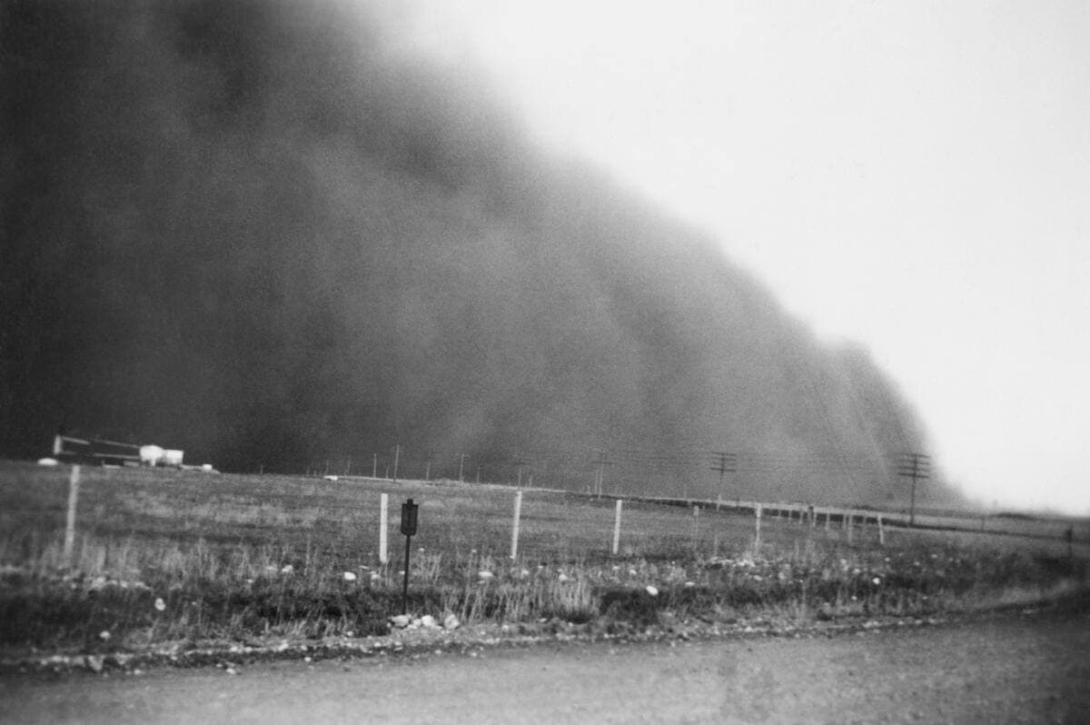 Black-and-white photograph of a towering wall of dust advancing over a flat field and a wire fence