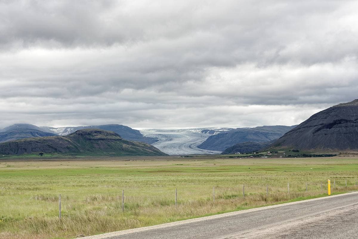 A distant glacier between mountains beyond flat green plain and a road