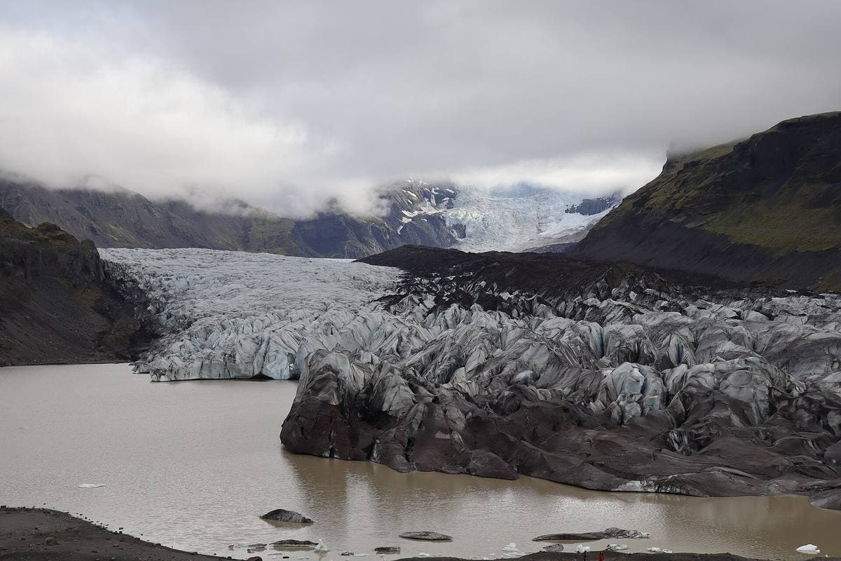 A grey and white glacier tongue descending between dark mountains into a still lagoon