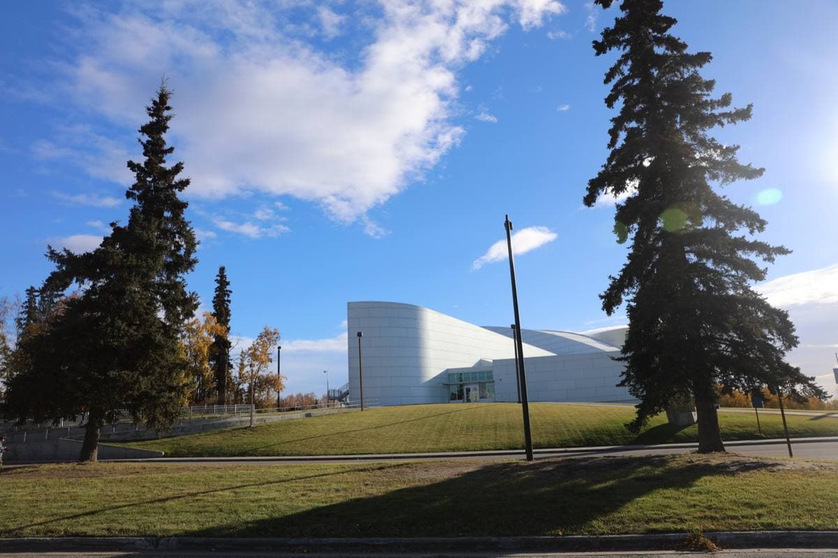 A white curving museum building seen across a lawn between tall spruces