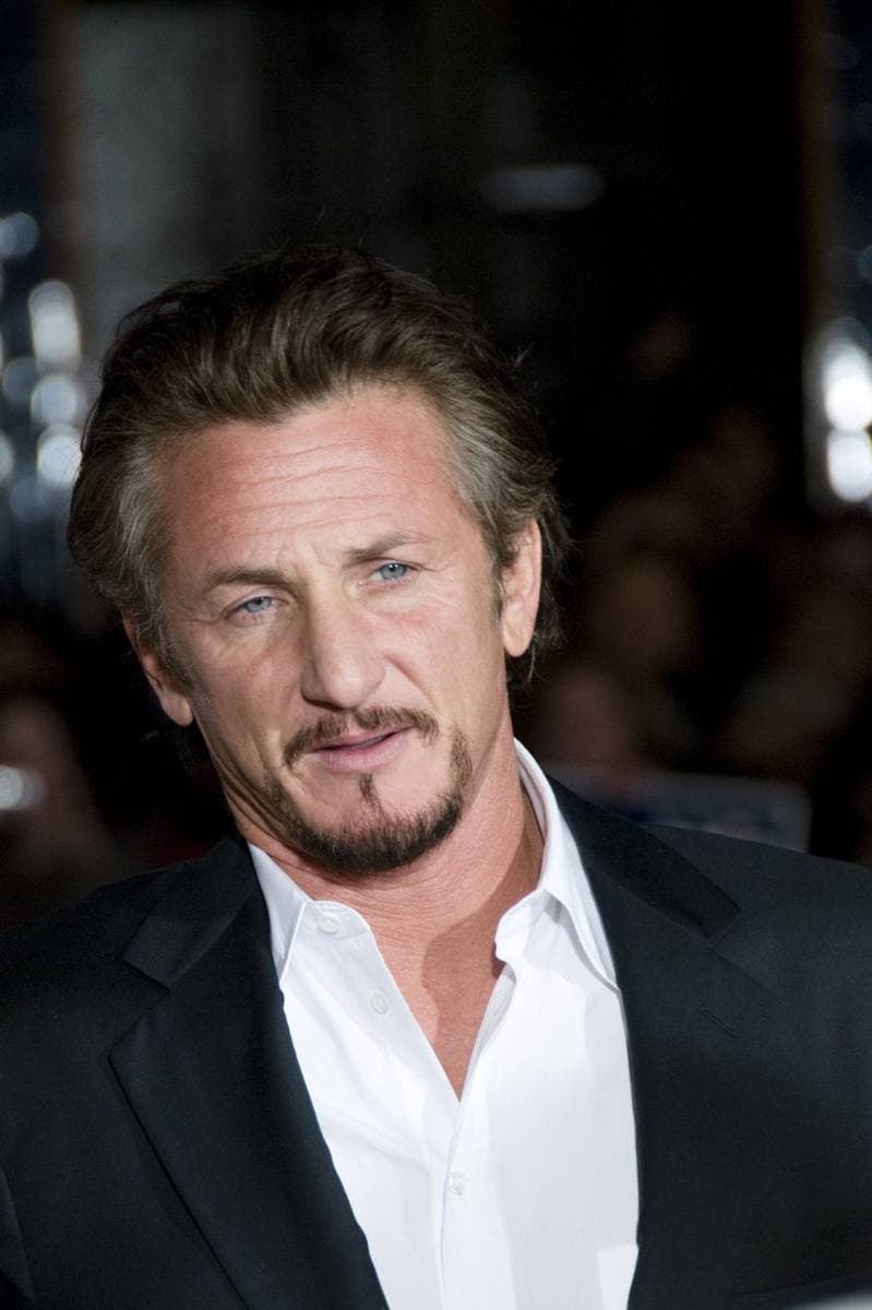 Sean Penn in a dark jacket and open white shirt, looking to one side