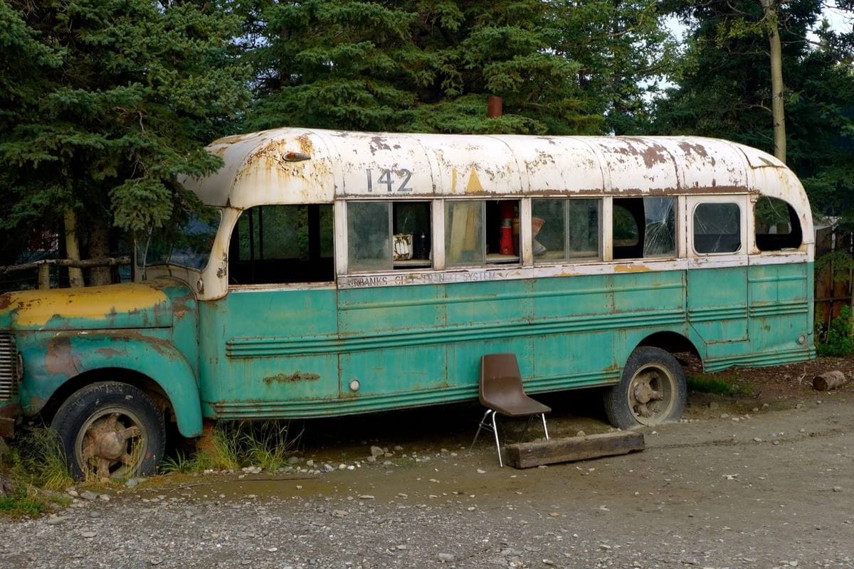 An old green-and-white bus with rusted wheel arches, parked among spruce trees