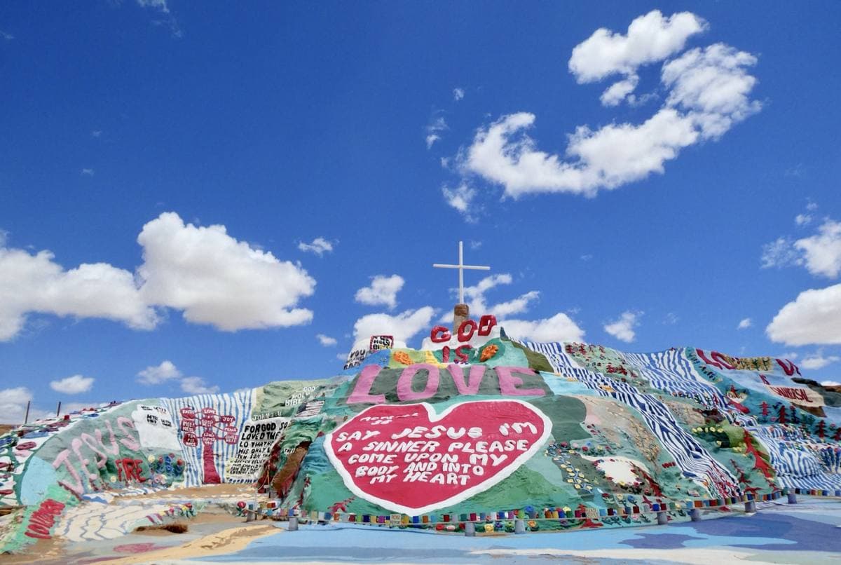 A hill painted in bright stripes and lettering under a blue sky with a white cross on top