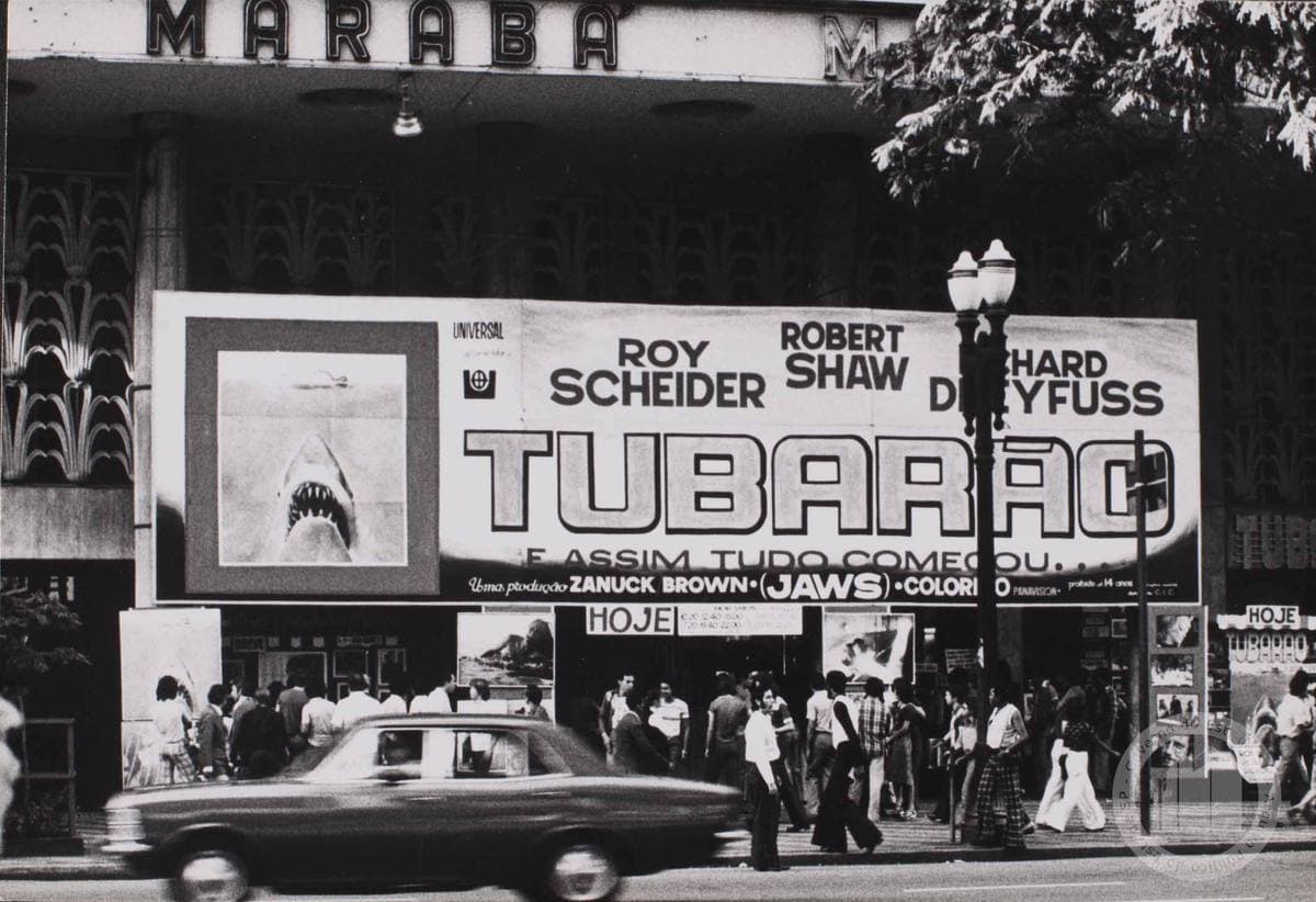 Black-and-white street photo of a cinema front with a huge Tubarão banner naming Roy Scheider, Robert Shaw and Richard Dreyfuss, a queue beneath and a car passing