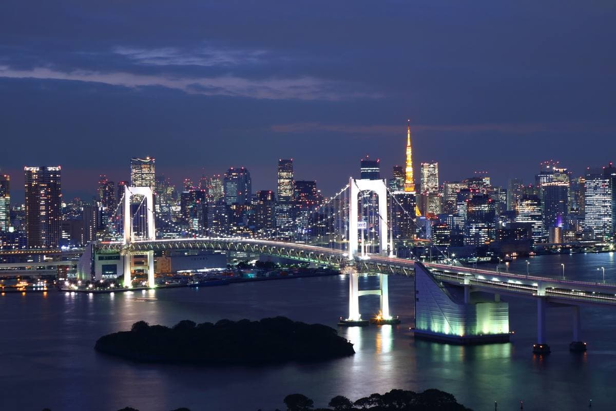 The Rainbow Bridge lit white over dark water, with the lit Tokyo skyline and Tokyo Tower behind