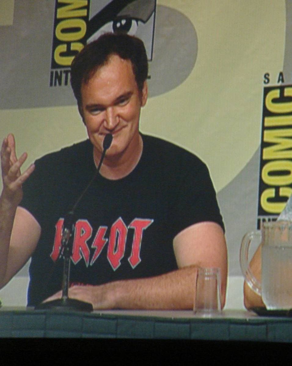 Quentin Tarantino in a black T-shirt, smiling at a panel microphone