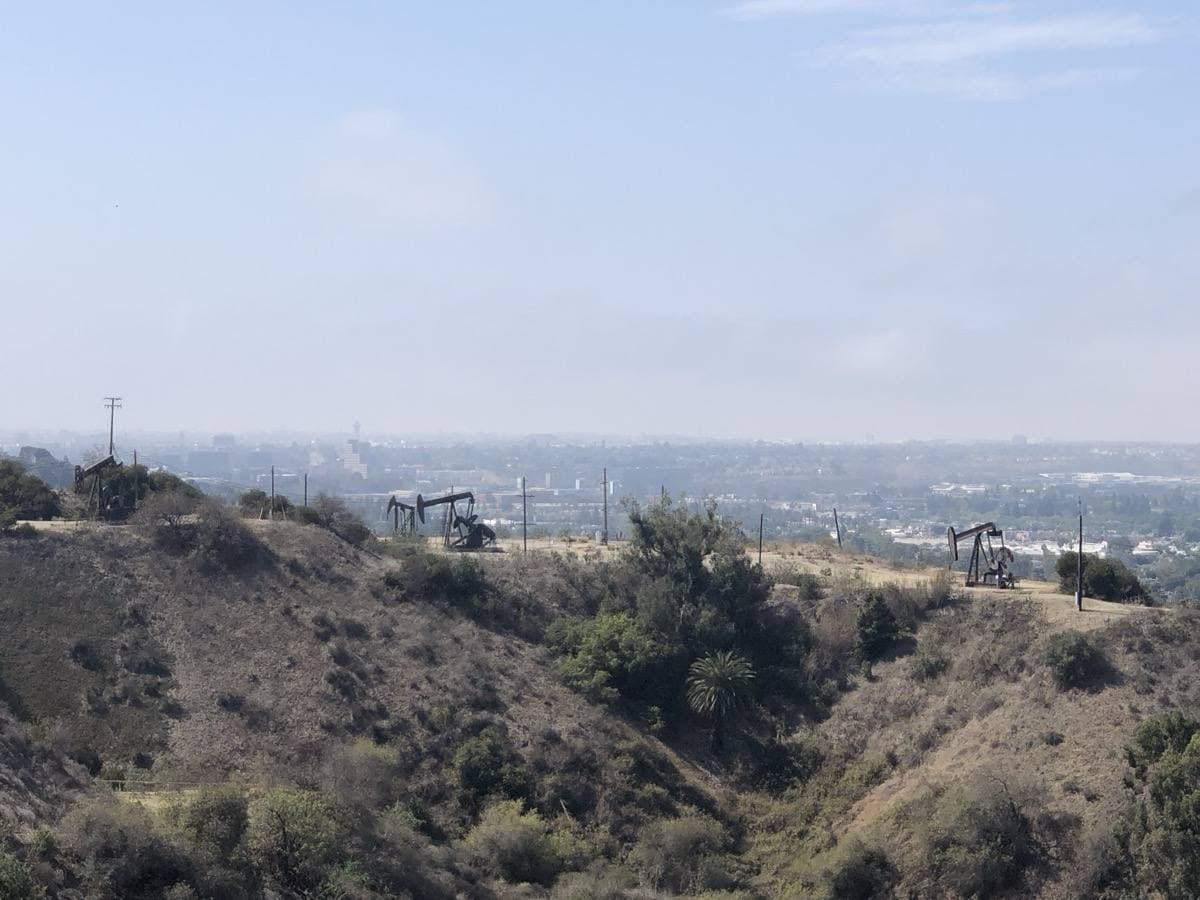 Three oil pumpjacks on dry scrubby hills, the city hazy beyond