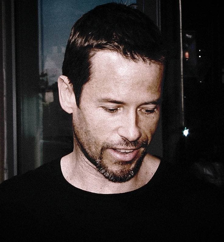 Guy Pearce with short dark hair and stubble, looking down and smiling