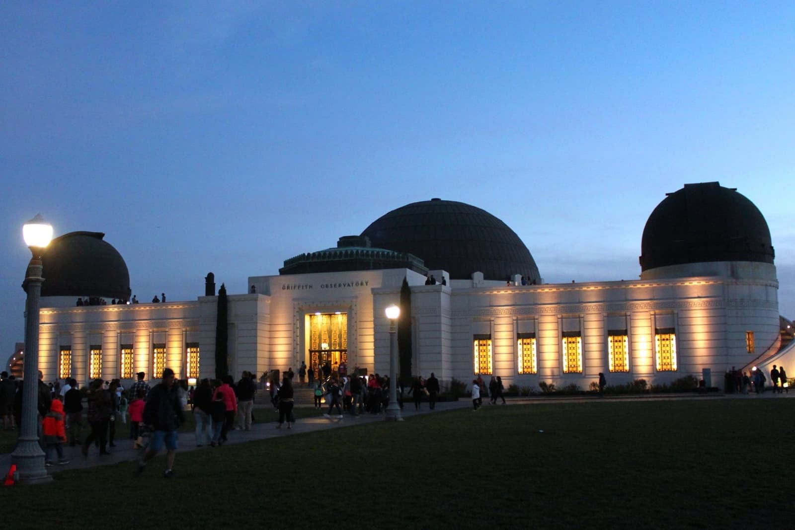 Griffith Observatory's white domes lit up at blue hour, with people on the lawn in front