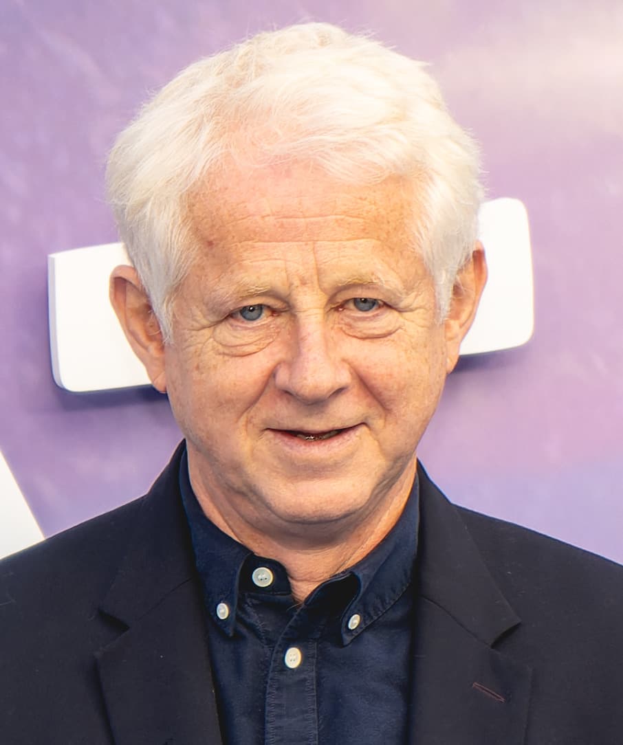 Richard Curtis, white-haired, smiling, in a dark jacket and blue shirt