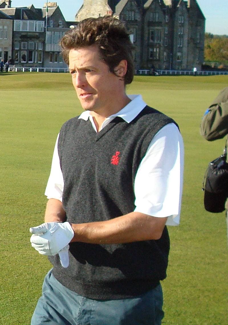 Hugh Grant in a grey sweater vest and white polo shirt on a golf course