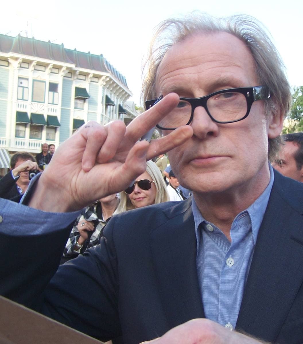 Bill Nighy in black-rimmed glasses and a dark suit, two fingers raised to his brow