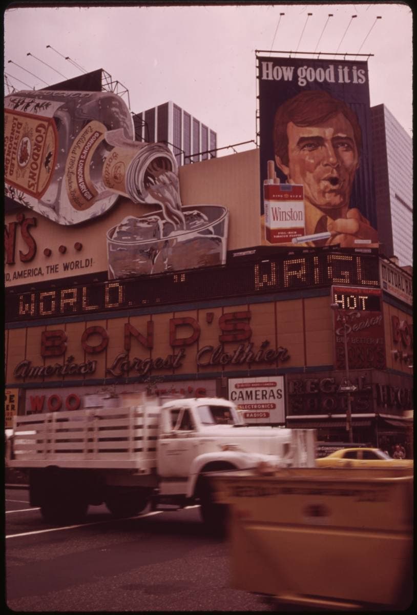 A Broadway storefront under billboards, a truck and a yellow cab passing