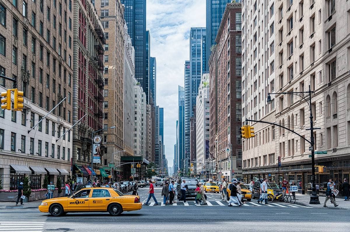 People crossing Sixth Avenue on a zebra crossing, a yellow cab waiting, towers behind