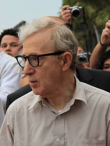 Woody Allen in black glasses and a beige shirt among a crowd of photographers
