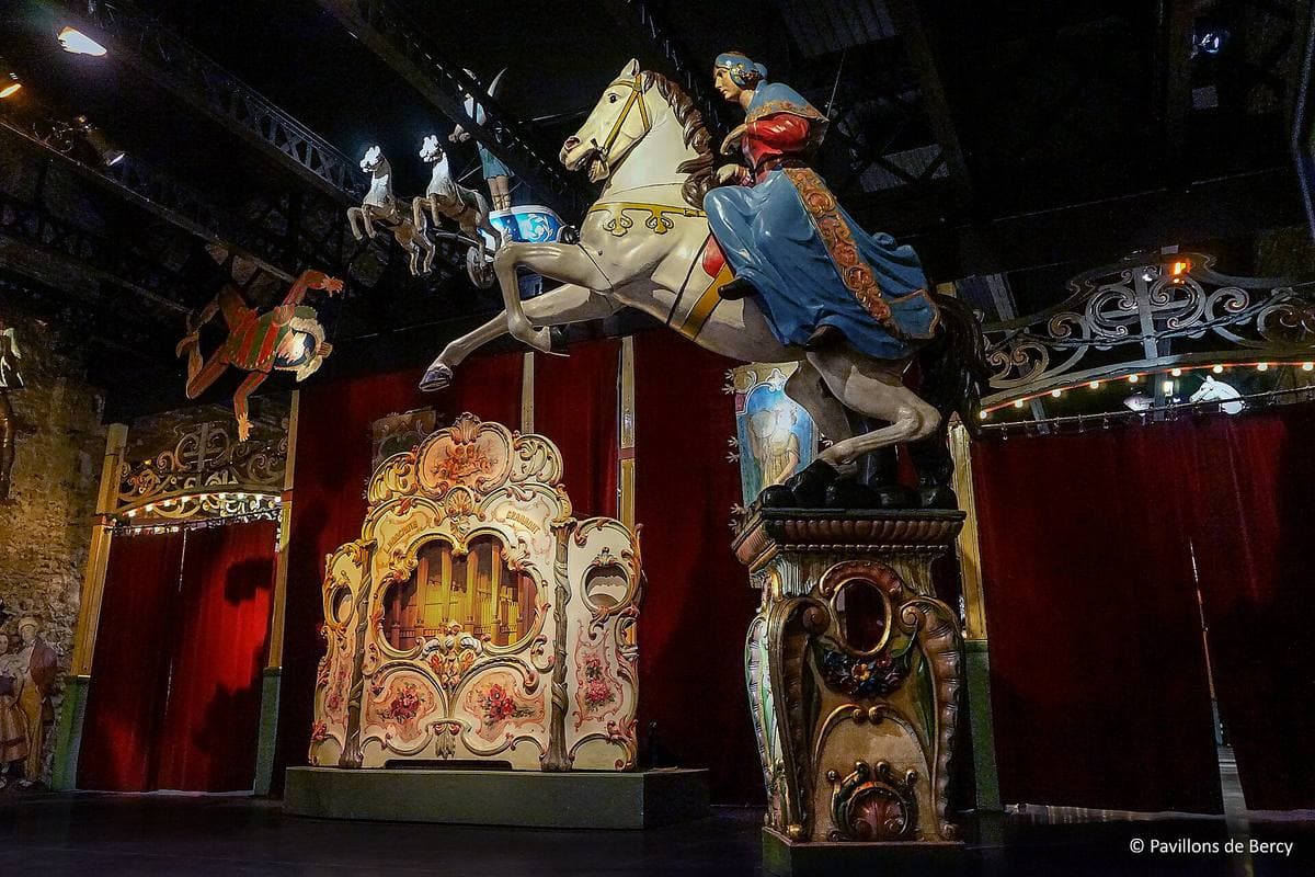 A hall of old fairground rides: a carved carousel horse and a gilded fair organ under red drapes