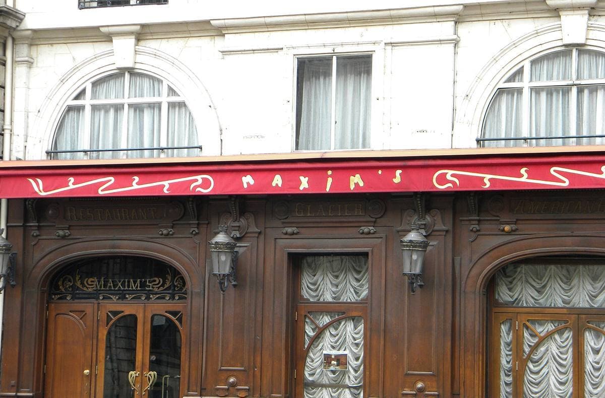 The carved dark-wood frontage of Maxim’s with a red awning lettered in gold and Art Nouveau lamps
