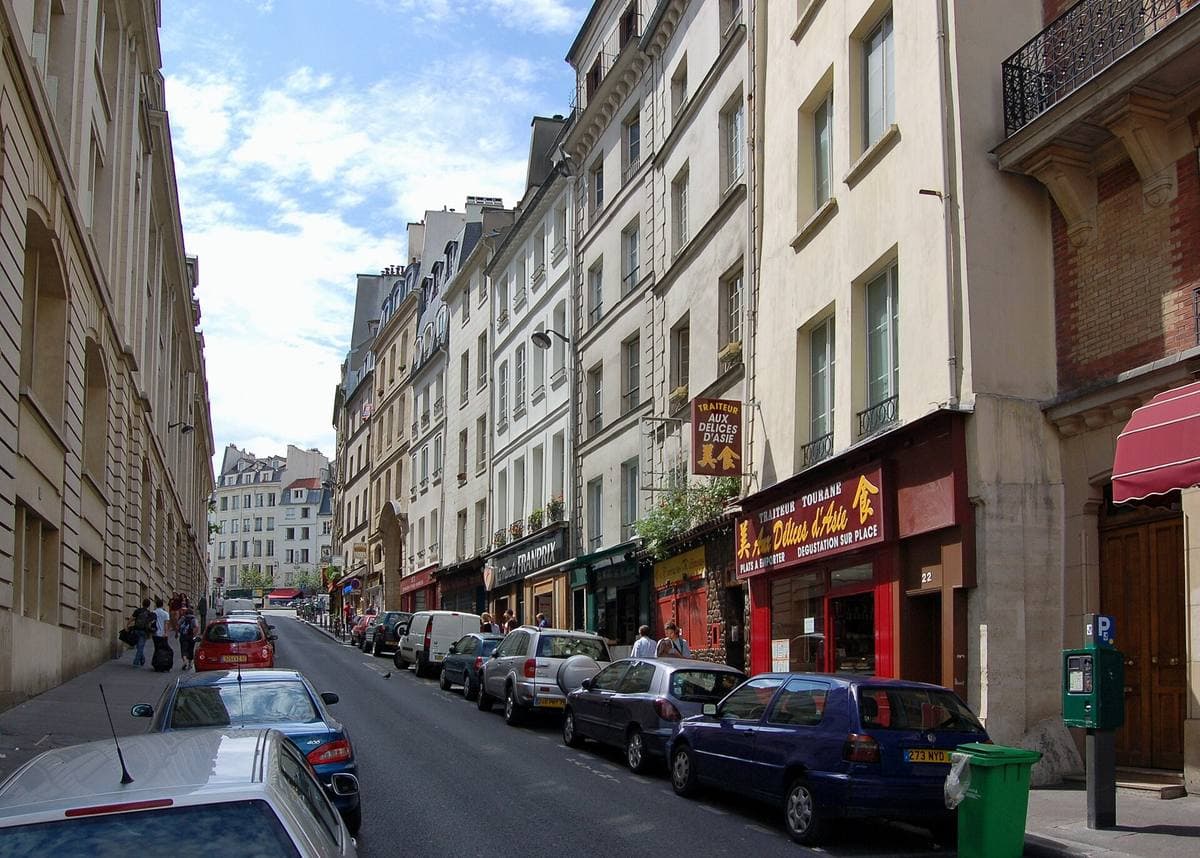 A narrow Paris street of tall pale buildings and parked cars running downhill
