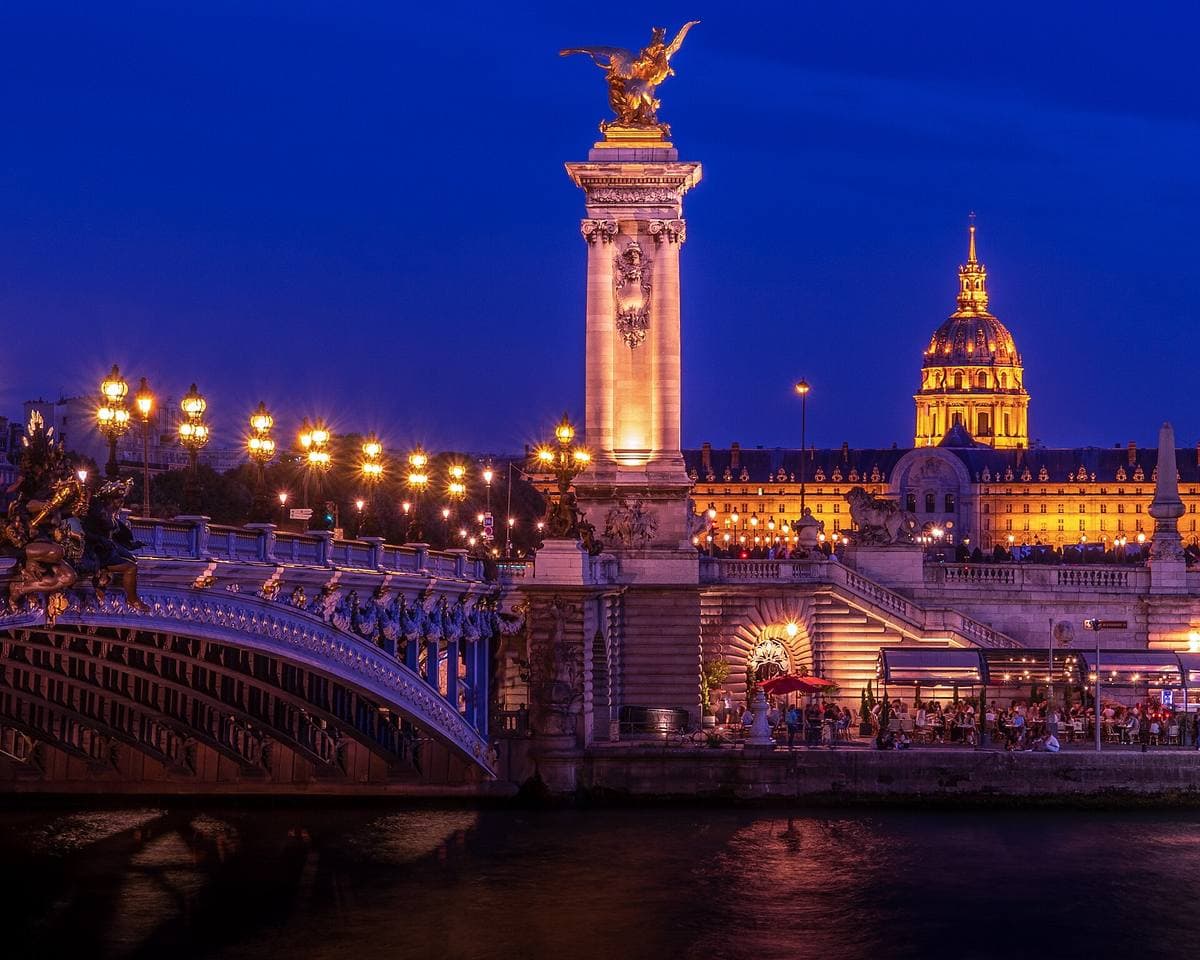 The Pont Alexandre III lit up at dusk with a gilded statue on its pillar and the golden dome of the Invalides behind