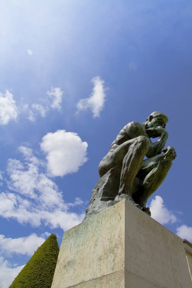 Rodin’s bronze Thinker on a tall stone plinth against a blue sky with white clouds