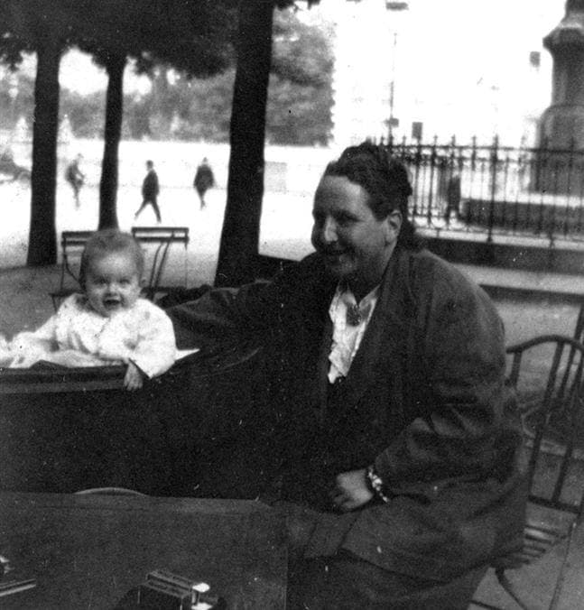 A black-and-white photograph of Gertrude Stein smiling at a baby in a pram in a Paris park