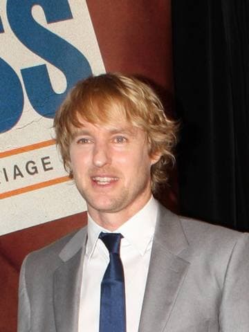 Owen Wilson smiling in a grey suit and blue tie