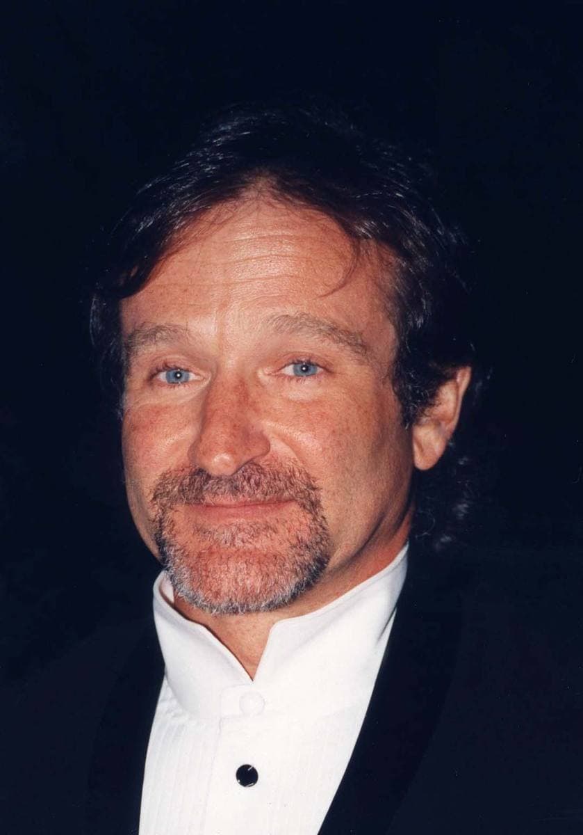 Robin Williams smiling, with a short greying beard, in a dark jacket and white collarless shirt