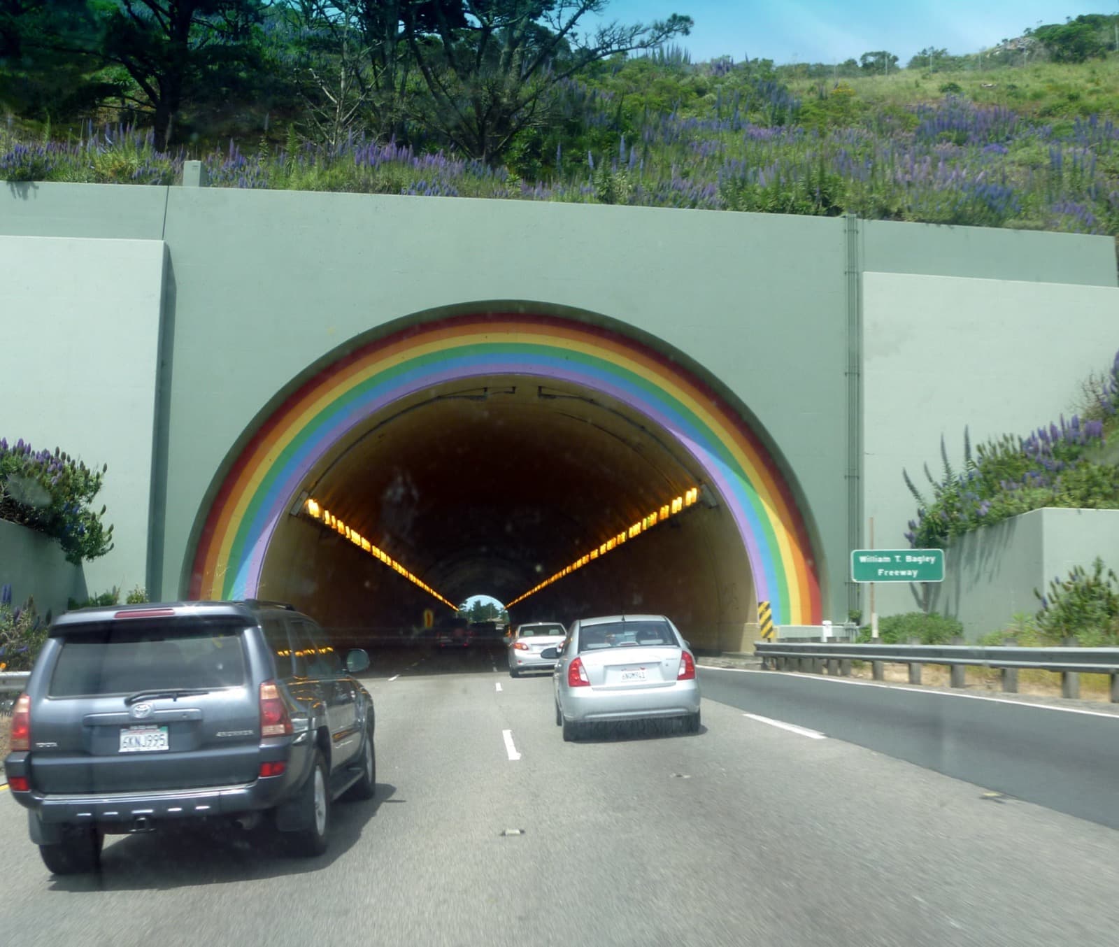 A highway tunnel mouth painted with a rainbow arch, cars driving in