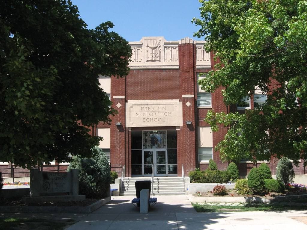 A red-brick school entrance with a carved stone panel above the doors, between two shade trees