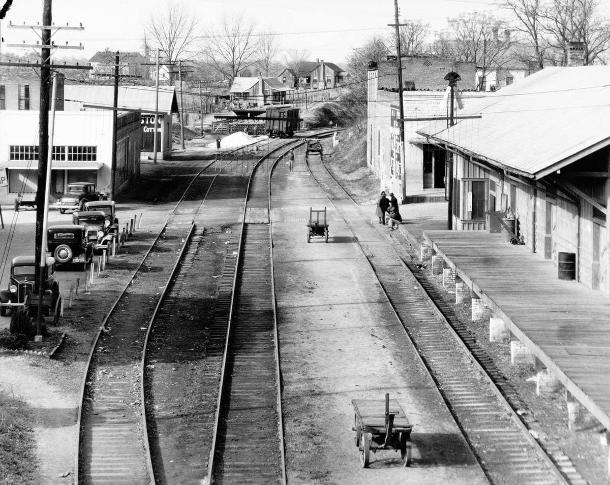 A black-and-white view down railroad tracks past a station and a small town