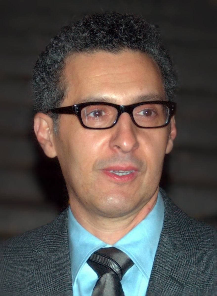 John Turturro in black-rimmed glasses, a pale blue shirt and grey jacket