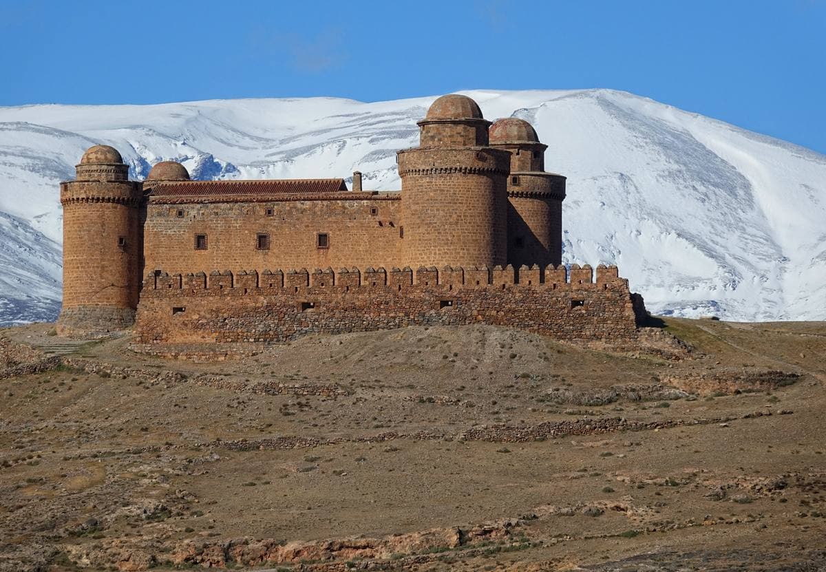 A square castle with round corner towers on a bare hill below snowy mountains