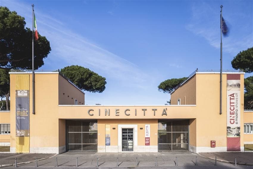 The pale yellow entrance building of Cinecittà with its name over the gate and two flagpoles