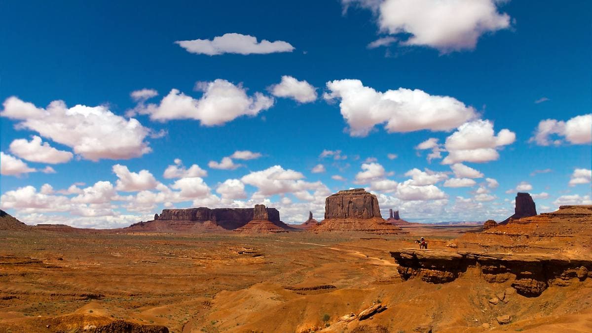 Red buttes on a wide plain under white clouds, seen from a rock ledge