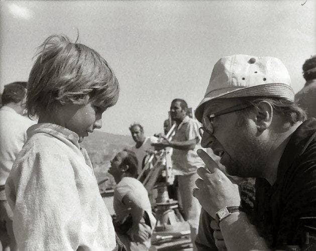 Sergio Leone in a white hat and glasses leaning towards a small boy on a sunlit set, crew behind