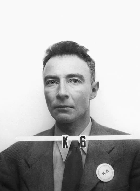 A black-and-white badge photograph of Oppenheimer facing the camera, a number card below his face